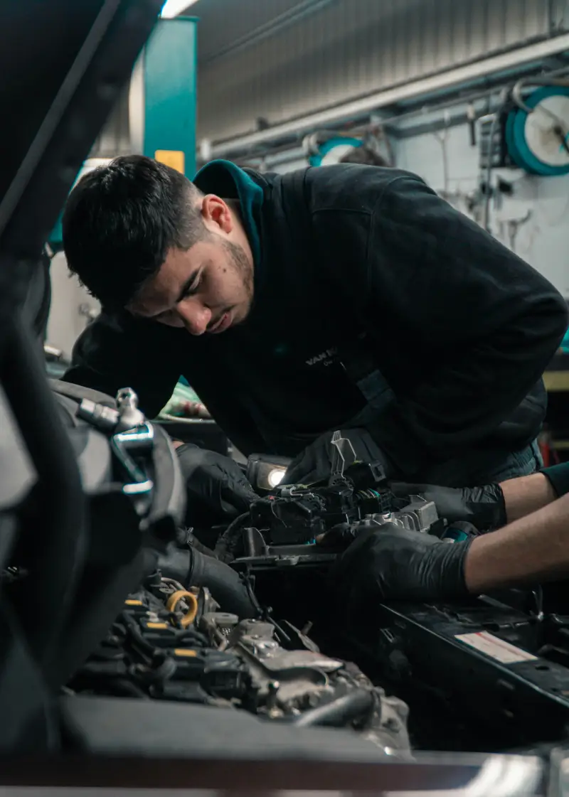 A mechanic in black gloves examines an engine component inside a car, focused and working in a well-equipped garage