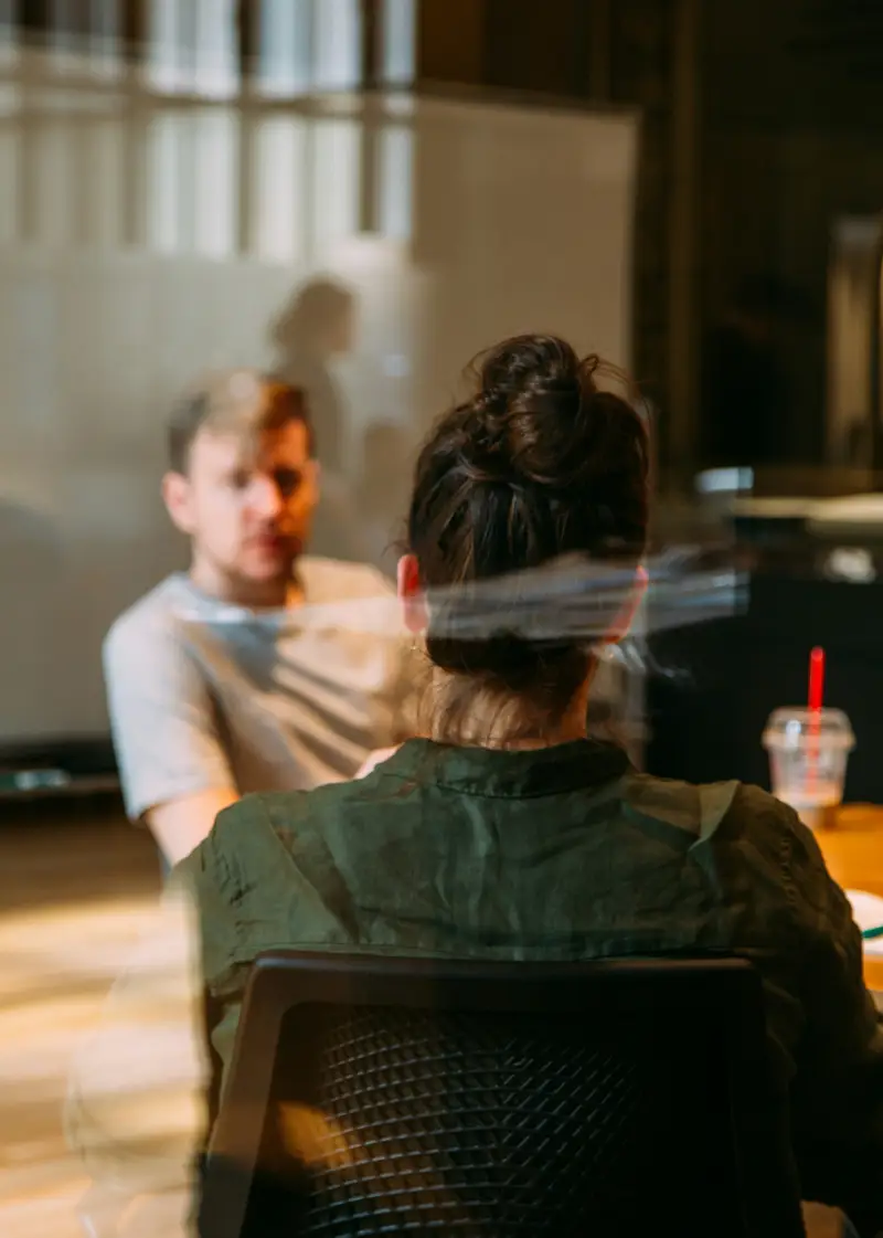 A woman with a bun hairstyle sits at a table in a meeting room, engaged in conversation, with a beverage beside her